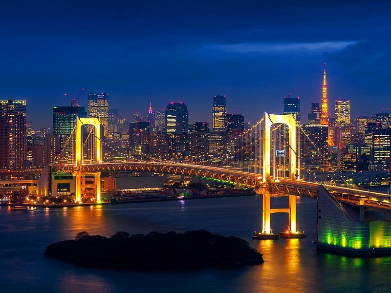 The bay bridge lit gold, the city towers and a red lattice tower behind it