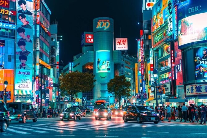A shopping street lit by neon signs and screens, taxis waiting at the lights