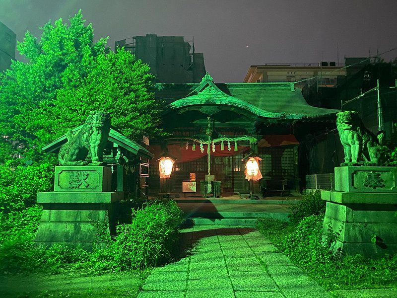 A small shrine at night, its stone guardians and path washed in green light